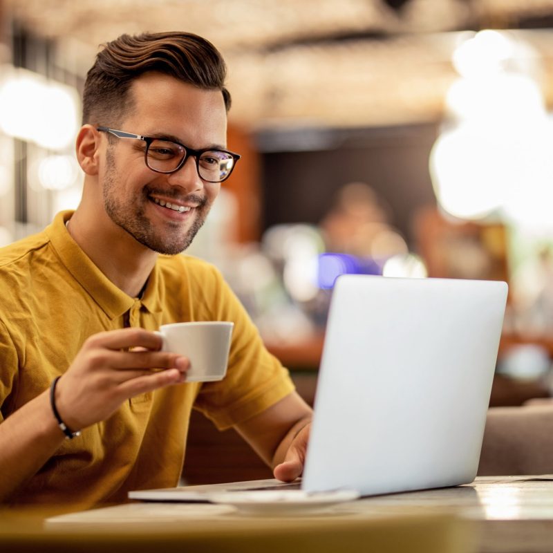 Smiling man surfing the net on laptop while drinking coffee in a bar.